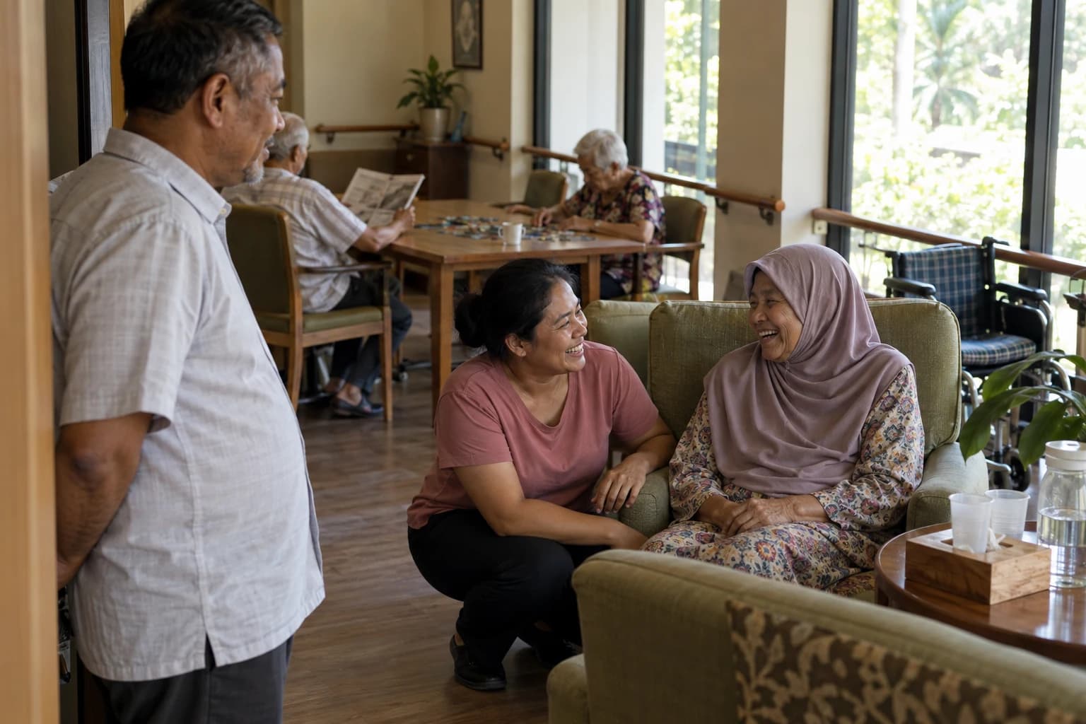 In a care home lounge, a care worker crouches to share a laugh with an older woman in a headscarf while a visiting man watches from the doorway