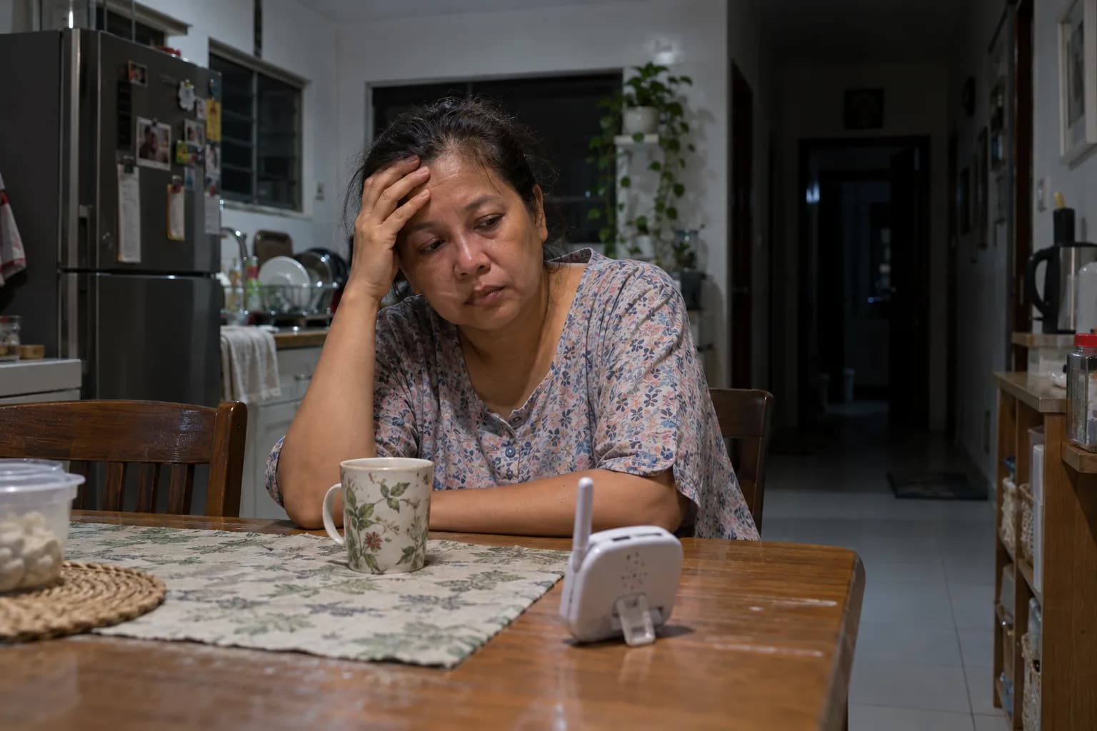 At night, a tired woman sits at the kitchen table with her head in her hand, a baby monitor beside her cup