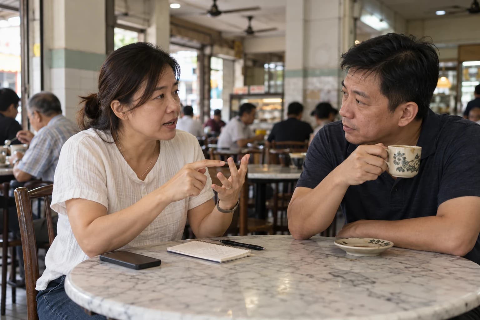 A brother and sister talk over coffee at a kopitiam table, one of them pointing at a notebook between them