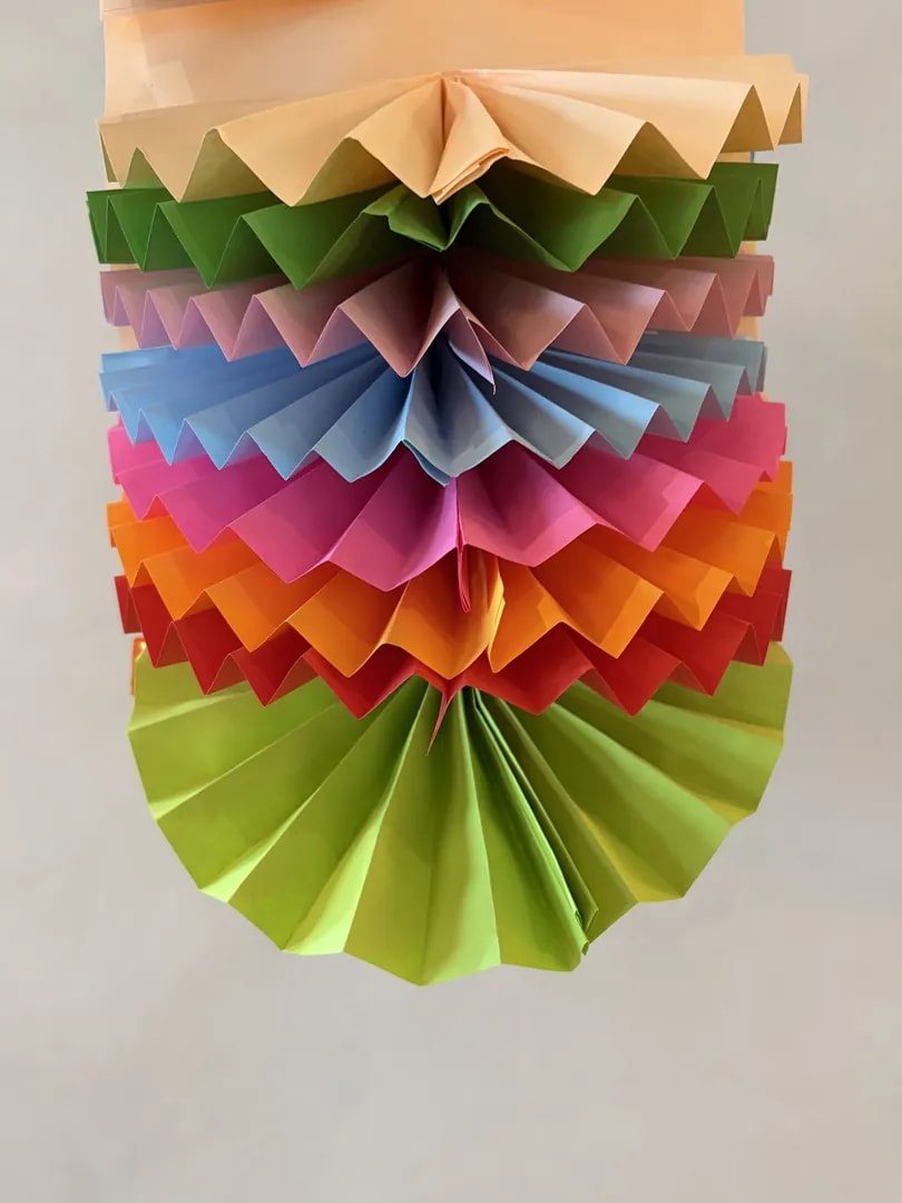 A folded paper lantern in bands of peach, green, pink, blue, orange, red and lime, hanging against a pale wall