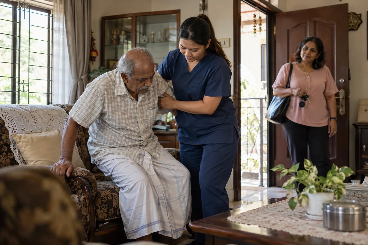 A caregiver in navy scrubs helps an older man rise from his armchair as his daughter picks up her bag to leave for work